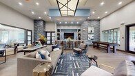 This single-floor community clubhouse is a thoughtfully designed gathering space that blends contemporary aesthetics with functional amenities. The open-concept layout is anchored by soaring ceilings and a striking geometric skylight feature overhead, flooding the lounge area with ambient light. Dramatic chevron-patterned textured wall panels in cool grey tones serve as a defining architectural element throughout, adding depth and visual interest to the space.

The main lounge area is furnished with plush, neutral-toned sofas and accent chairs arranged over a bold navy and white geometric area rug, creating an inviting setting for socializing. Wide-plank wood flooring flows throughout, unifying the various zones seamlessly. A central bar-height counter with warm wood accents and bar seating connects the lounge to an entertainment wall featuring a large flat-screen TV flanked by open display shelving with light blue insets.

Adjacent to the entertainment area, a shuffleboard table provides a recreational focal point, while curated abstract artwork adorns the walls nearby. A dedicated private room, accessible through glass-paneled doors and featuring a decorative textured glass window, offers a more intimate setting with cowhide-accented seating. Near the entry, a casual dining nook with round tables and upholstered chairs provides an additional gathering option alongside a built-in service station.