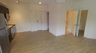 This single-floor unit presents a clean, modern aesthetic defined by its open layout and neutral palette. The main living area is finished with light-toned hardwood-style flooring that runs throughout, lending warmth and continuity to the space. Bright white walls reflect available light, contributing to an airy, well-lit atmosphere.

At the far end of the main area, a white paneled entry door is equipped with a keypad lock, offering both security and convenience. Adjacent to the entry is what appears to be a closet, providing practical storage within easy reach of the entrance. A partial wall creates a subtle division within the open floor plan, adding a degree of structural definition without enclosing the space.

To the right, a doorway opens into a bathroom, where fixtures are partially visible, suggesting a functional and neatly appointed layout. Flush-mounted ceiling lighting provides consistent illumination throughout. The overall design is straightforward and functional, with a move-in-ready condition that offers a neutral foundation well-suited for a variety of interior styles.