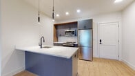 This single-floor residence at 20 Grand showcases a thoughtfully designed contemporary interior with clean lines and modern finishes throughout. The kitchen serves as a focal point, centered around a striking island with a sleek blue base and veined white quartz countertop, illuminated by cylindrical pendant lights. Matte gray cabinetry is framed by warm wood accents, complemented by a white subway tile backsplash and a full suite of stainless steel appliances, including a refrigerator, range, and microwave. Light hardwood flooring flows seamlessly through the open living areas, creating a warm and cohesive aesthetic.

The bedroom features a window with an exterior view, allowing natural light to fill the space, along with a wall-mounted HVAC unit for individual climate control. Two bathrooms serve the unit — one with a floating wood vanity, backlit mirror, and gray tile flooring, and a second more utilitarian space finished in the same neutral tile palette. The entry door is equipped with a smart lock, adding a practical modern touch. Throughout, the unit maintains a consistent palette of warm whites, natural wood tones, and cool grays, resulting in a clean, move-in-ready environment.