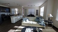 This stylish single-floor residence showcases a thoughtfully designed open-concept layout that effortlessly blends the living and kitchen areas into one cohesive, inviting space. The living area is anchored by a plush, cream-toned sofa accented with soft blue and gray throw pillows, complemented by a sleek dark coffee table and a cozy area rug. Multiple windows fitted with white blinds allow generous natural light to flood the space, creating a bright and airy ambiance throughout the day. Track lighting on the ceiling provides additional illumination, enhancing the clean, contemporary aesthetic. The kitchen is appointed with crisp white cabinetry, granite countertops, and a peninsula-style breakfast bar lined with modern white bar stools — ideal for casual dining or entertaining. Pendant lights hang elegantly above the bar, adding a warm, refined touch. Dark hardwood-style flooring runs throughout, providing a striking contrast to the light-colored furnishings and walls. Decorative wall art and tasteful accents add personality to the space, while the overall design maintains a cohesive, modern sensibility. A glass-panel door leads to an exterior entry, further enhancing the sense of openness and connectivity in this well-appointed home.