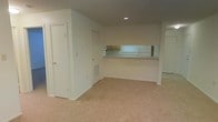 This single-floor space offers a clean, functional layout designed for comfortable everyday living. The main living area is finished with wall-to-wall carpeting in a neutral beige tone, providing a warm and quiet underfoot feel throughout the space. Recessed lighting in the ceiling provides focused illumination, complementing the bright white walls that keep the interior feeling open and airy.

A defining architectural feature is the pass-through counter that connects the kitchen to the living area. This opening allows natural light from the kitchen's window to filter into the main room while creating a sense of connectivity between the two spaces. The kitchen, visible through the pass-through, includes upper cabinetry in a warm wood finish and appears well-organized and efficiently laid out.

Multiple doorways lead to additional areas of the unit, suggesting the presence of closet or utility storage alongside other rooms. A floor-level HVAC vent is integrated into the wall, indicating a central air and heating system. The overall design is straightforward and practical, with white walls, clean lines, and a neutral palette that offers a versatile canvas for a variety of interior styles. The space is well-suited for those seeking a tidy, move-in ready environment with an efficient, connected floor plan.