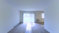 This single-floor space features an open living area finished with wall-to-wall carpeting and crisp white walls that contribute to a clean, airy atmosphere. The room is anchored by a large sliding glass door fitted with vertical blinds, which serves as the primary source of natural light and provides a direct view of a green outdoor area. An additional window on the adjacent wall further brightens the space, ensuring consistent illumination throughout the day. A baseboard heating unit runs along the lower portion of the exterior wall, offering an efficient heating solution, while a wall-mounted air conditioning unit provides climate control during warmer months. The layout of the living area is straightforward and open, lending itself well to flexible furniture arrangements. The white ceiling and neutral-toned carpet create a cohesive, understated aesthetic. Overall, the space presents a functional and well-lit environment with practical climate control features and easy access to the outdoors through its prominent sliding glass door.