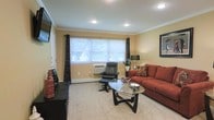 This single-floor space presents a thoughtfully arranged living area that balances comfort and functionality. The living room is finished with warm yellow-toned walls accented by white crown molding, creating a clean and inviting atmosphere. Recessed ceiling lights provide consistent, even illumination throughout the room, complementing the natural light that filters in through a wide, centrally positioned window dressed with patterned drapes and horizontal blinds. The window offers views of surrounding greenery, adding a pleasant outdoor connection to the interior.

The room features light-toned carpeting underfoot, contributing to its warm and relaxed ambiance. A rich, rust-colored sofa serves as the focal point of the seating area, paired with a black leather recliner and ottoman for additional comfort. A contemporary oval glass coffee table anchors the arrangement, while a dark wood media console along one wall provides practical storage and display space. A wall-mounted television adds a modern touch. The space is further accented by a floor lamp, decorative accessories, and framed artwork, rounding out the room's cohesive and livable character.