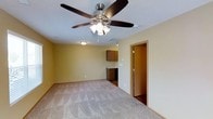 This single-floor space offers a straightforward, functional layout designed for comfortable everyday living. The main living area is finished with wall-to-wall carpeting in a neutral tone, providing a soft underfoot feel that complements the warm yellow-beige walls throughout. A prominent ceiling fan with an integrated multi-bulb light fixture is centrally mounted, ensuring both air circulation and ambient lighting across the open space.

Toward the back of the main area, a compact kitchen zone is visible, featuring medium-toned wood cabinetry mounted above a countertop with a dark appliance below — likely a range or dishwasher. The kitchen area transitions to a hard flooring surface, creating a practical distinction between the living and cooking zones.

An interior doorway along the right wall leads to an adjoining room, suggesting additional functional space such as a bedroom or utility area. The transition between rooms features a darker flooring material, adding subtle contrast. Additional flush-mount lighting in the hallway area ensures the connected spaces remain well-lit. Overall, the space presents a clean, no-frills interior suitable for efficient, day-to-day use.