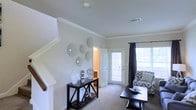 This two-story home offers a thoughtfully designed layout that balances comfort and functionality. The main living area greets visitors with a bright, airy atmosphere, thanks to large windows dressed with dark curtain panels that allow abundant natural light to filter in. Soft, neutral wall tones are complemented by plush carpeting underfoot, creating a warm and inviting environment throughout.

Crown molding adds a refined architectural touch to the living space, which is accented by decorative wall art and a stylish console table — details that lend the room a curated, polished feel. A glass-paneled front door further enhances the sense of openness while connecting the interior to the outdoors. A staircase leads to the upper floor, offering additional living or sleeping spaces above.

A doorway visible from the living area hints at additional rooms, suggesting a practical floor plan with well-defined zones for living, storage, and utility. The overall ambiance of the home is clean, neutral, and move-in ready, with tasteful decor choices that highlight the space's inherent architectural qualities. The combination of crown molding, natural light, and soft finishes makes this a comfortable and visually cohesive two-story residence.