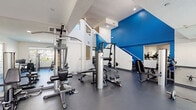 This two-story fitness center, located in the San Francisco Bay Area (94597), offers a comprehensive workout facility designed to accommodate a wide range of exercise needs. The space is organized across two floors, each serving distinct training purposes, connected by an interior staircase.

The lower level is anchored by a spacious strength training area featuring multiple cable machines, weight benches, free weights, and a TuffStuff functional trainer. A bold blue accent wall adds a vibrant, energizing aesthetic to the space, complemented by white walls, high ceilings, and durable rubber flooring throughout. Large windows allow natural light to flow in, creating a bright and open environment.

Also on the lower level is a dedicated barre and stretching zone, outfitted with floor-to-ceiling mirrors, a wall-mounted wooden barre rail, and open floor space ideal for flexibility and low-impact training.

The upper level houses a cardio-focused area equipped with multiple elliptical trainers, treadmills, and a rowing machine. Full-length mirrors line the walls, enhancing the sense of space, while recessed lighting and windows overlooking the surrounding greenery keep the area bright and inviting.

Together, the two floors create a well-rounded fitness facility that supports strength, cardio, and functional training in a clean, modern, and thoughtfully designed setting.