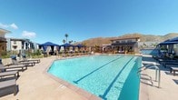 This single-story amenity facility is thoughtfully designed to support health and recreation. The centerpiece of the outdoor area is a generously sized swimming pool with crystal-clear blue water, surrounded by a spacious sun deck furnished with lounge chairs and private cabanas draped in navy blue fabric. Palm trees and desert landscaping frame the space, while dramatic mountain views provide a striking natural backdrop beneath open skies.

The interior fitness center is architecturally notable for its soaring ceilings, enhanced by clerestory windows that line the upper walls and flood the space with natural light. A large ceiling fan provides air circulation throughout the open workout floor. The facility is outfitted with a comprehensive selection of Technogym cardio equipment, including treadmills, ellipticals, and rowing machines, all positioned along floor-to-ceiling windows overlooking the outdoors. Strength training equipment, cable machines, free weights, and resistance stations are arranged throughout the mirrored main floor.

A front check-in counter with built-in storage cubbies greets users upon entry. The facility also includes a dedicated sanitation station with a built-in sink, dark wood cabinetry, white countertops, and stainless steel waste disposal, ensuring cleanliness and convenience throughout.