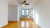 This single-floor residence offers a well-organized layout that balances open living space with functional utility. The main living area is immediately inviting, characterized by warm, polished hardwood floors that run throughout and crisp white walls that amplify the abundant natural light pouring in through multiple black-framed double-hung windows. A ceiling fan with an integrated light fixture serves as a central feature overhead. Built-in shelving flanks a radiator beneath the windows, providing both practical storage and a charming architectural detail. The panoramic city views visible from multiple windows give the space a notably elevated and airy feel.

The galley-style kitchen is efficiently designed, featuring warm oak cabinetry with ample upper and lower storage, a double-basin stainless steel sink, granite-look countertops, and white appliances including a stove and refrigerator. Light ceramic tile flooring completes the practical aesthetic of this workspace.

The bathroom is a classic highlight, finished in timeless black-and-white octagonal mosaic tile flooring, white subway-tiled walls with dark navy accent trim, and a full soaking bathtub. A frosted window provides natural light while maintaining privacy.

Throughout the space, the consistent use of hardwood floors, neutral walls, and traditional architectural details create a cohesive and comfortable living environment with strong urban character.