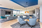 This contemporary amenity lounge is a thoughtfully designed social space spread across a single floor, combining vibrant aesthetics with functional versatility. The interior is defined by striking cobalt blue accent walls and ceiling panels that contrast boldly against crisp white surfaces, creating an energetic and modern atmosphere throughout.

The main open-plan area serves multiple functions seamlessly. A comfortable lounge zone features a grey sectional sofa, an assortment of swivel chairs in shades of blue and teal, and area rugs anchoring each seating cluster. A wall-mounted flat-screen television and a textured geometric accent wall serve as a focal point for the media area.

A dedicated recreation zone houses a full-size ping-pong table positioned near expansive floor-to-ceiling windows that flood the space with natural light and offer views of the surrounding neighborhood. Adjacent to this is a sleek kitchenette with white cabinetry, a countertop workspace, and bar seating with teal cushioned stools.

A private meeting or dining nook, enclosed in glass, features pendant lighting with warm metallic tones, providing a more intimate setting within the larger open floor plan. Light wood-toned flooring runs throughout, lending warmth and cohesion to the space.

Large bi-fold glass doors open directly onto an outdoor terrace, extending the living and entertaining area outside and allowing for abundant natural light and seamless indoor-outdoor flow.