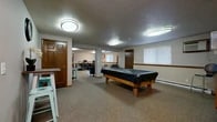 The clubhouse at 1905 W Appleway Ave in Coeur d'Alene, Idaho, is a single-floor community amenity space designed to serve a variety of recreational and practical needs. The layout flows across several distinct areas, each carpeted or finished with durable flooring suited to its function.

The main recreation area is centered around a full-size pool table and is accented with warm wood-trimmed doors and neutral-toned walls. Adjacent to the billiards area is a comfortable lounge featuring a wall-mounted flat-screen television, a small entertainment console, upholstered seating, and bar-height tables with teal metal stools that add a pop of color to the space.

A dedicated business center area provides multiple computer workstations along a long desk, accompanied by bar stools, a printer, and a cozy seating nook with a leather sofa and accent chair set atop a decorative area rug—ideal for residents needing a quiet workspace.

The fitness room is well-appointed with a multi-station cable weight machine, a treadmill, a stationary bike, and a rack of medicine balls positioned beside a teal accent wall, offering a motivating environment for exercise.

Throughout the clubhouse, textured ceilings, flush-mount lighting, and windows with blinds contribute to a clean, functional atmosphere that balances community gathering with everyday utility.