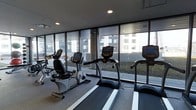 This single-floor fitness center offers a clean, contemporary workout environment designed to support an active lifestyle. The space is outfitted with a selection of high-quality True-brand cardio equipment, including treadmills, upright bikes, and a recumbent bike, providing users with a variety of cardiovascular training options. Each machine features an integrated digital display, enabling users to track performance metrics with ease. The flooring is finished with durable, textured fitness flooring suitable for high-traffic use. One of the most notable features of this gym is the expansive floor-to-ceiling window wall that lines the perimeter, flooding the space with abundant natural light and offering open views of the surrounding outdoor terrace and adjacent buildings. This connection to the outdoors creates an airy, motivating atmosphere that enhances the overall workout experience. Recessed ceiling lighting supplements the natural light, ensuring the space remains well-lit at all hours. The layout is thoughtfully arranged to allow comfortable spacing between equipment, promoting ease of movement throughout the room. Overall, the fitness center combines functional design with a bright, modern aesthetic.