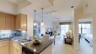 This single-floor residence features an open-concept layout that seamlessly connects the kitchen, dining, and living areas, creating a cohesive and functional living environment. The kitchen is appointed with dark granite countertops, a double stainless steel sink, warm wood cabinetry, and a tiled backsplash. A central island provides additional workspace and casual dining opportunities, while pendant lighting and a track lighting system illuminate the space effectively.

The dining area sits adjacent to the kitchen, defined by its own overhead pendant light and furnished with a round table and upholstered chairs. Beyond the dining space, the living room offers a comfortable, well-lit environment with neutral tones, a ceiling fan, and a wall-mounted television. Large sliding glass doors at the far end of the living room flood the interior with natural light and open onto a private balcony, offering views of the surrounding area. The overall ambiance is modern and welcoming, with a consistent neutral palette, light flooring, and carefully placed décor throughout.