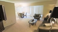 This single-floor residence features an open-concept layout that seamlessly connects the living and dining areas, creating a cohesive and functional living environment. The living area is carpeted in a neutral, warm tone that extends throughout the space, contributing to a soft and inviting atmosphere. A glass-topped oval coffee table serves as a focal point in the living area, complemented by upholstered seating including a patterned accent chair and a sofa. The dining area, positioned adjacent to the living space, accommodates a rectangular table with a set of high-backed upholstered chairs beneath a pendant light fixture. Large vertical-blind-covered windows and a sliding glass door line the far wall, allowing generous natural light to fill the space while offering access to the outdoors. The neutral wall tones and light color palette throughout reinforce a clean, understated aesthetic. A framed piece of wall art adds a decorative accent near the dining area. Overall, the open layout, plentiful natural light, and consistent flooring create a cohesive and comfortable living environment.