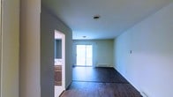 This single-floor space is defined by its clean, open layout and straightforward design. The main living area is finished with rich, dark hardwood flooring that contrasts with the light-toned walls, creating a simple yet appealing aesthetic. A sliding glass door anchors the far wall of the living area, flooding the room with natural light and providing direct access to the outdoors. Beneath the window, a baseboard heating unit ensures comfort throughout the seasons. The ceiling is smooth and white, fitted with a sprinkler head, indicating a modern safety system within the building. To one side, an open doorway leads directly into a bathroom, which features warm wood-toned cabinetry and a sink, offering a functional and accessible layout. The walls throughout are painted in a neutral, muted tone that keeps the space feeling calm and versatile. The overall ambiance is understated and practical, offering a blank canvas well-suited for a variety of interior arrangements. The single-floor configuration ensures easy accessibility and a cohesive flow throughout the unit.