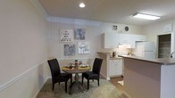 This single-floor residence features an open-concept layout that seamlessly connects the dining area and kitchen, creating a cohesive and functional living space. The dining area is intimate and inviting, centered around a round wooden pedestal table paired with two dark upholstered chairs, set atop a textured area rug. Decorative wall art, including motivational typography pieces in neutral tones, adds a personal and cheerful character to the space. A chair rail detail runs along the lower portion of the walls, lending a classic architectural touch.

The adjacent kitchen is well-appointed with white cabinetry offering ample storage, light countertops, and white appliances including a range. An under-cabinet light fixture provides task lighting over the workspace. The kitchen transitions smoothly into the dining space, supported by a partial dividing counter that defines each zone without closing off the open feel. Recessed lighting in the dining area and overhead fluorescent lighting in the kitchen ensure the space is well-lit throughout. Crown molding along the ceiling adds a refined finishing detail. The overall ambiance is warm, tidy, and thoughtfully arranged.