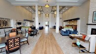 The Orchards Arlington Heights community clubhouse at 131 E Bardin Rd in Arlington, Texas, serves as a welcoming social hub for this active adult community. The single-floor space is defined by soaring ceilings and an expansive open-plan layout anchored by striking white stacked-stone columns that rise dramatically toward the ceiling, lending a refined yet approachable character to the interior.

The great room is divided into distinct yet flowing zones. A comfortable lounge area features a floor-to-ceiling stacked-stone fireplace flanked by plush blue velvet armchairs and a neutral sofa with accent pillows, creating an inviting gathering spot. Adjacent to this is a well-appointed dining area with dark-wood tables and chairs with orange and white upholstery, framed by decorative tree-motif partitions that add an artistic touch.

A kitchenette bar with dark cabinetry and a mounted television provides convenient service space for events and daily use. Hardwood flooring runs along the central corridor, transitioning to patterned carpet in the seating and dining zones. Recessed lighting and pendant fixtures illuminate the space throughout.

Large floor-to-ceiling windows and glass doors along one wall flood the interior with natural light and offer direct views of the outdoor pool courtyard with patio seating and umbrellas. Decorative wall accents and curated artwork complete the polished, community-focused ambiance.