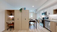 This efficiently designed single-floor studio makes excellent use of its footprint through smart layout planning and contemporary finishes. The open-plan kitchen and living area forms the heart of the space, featuring a fully equipped kitchen with stainless steel appliances — including a refrigerator, range, dishwasher, and microwave — complemented by white upper cabinetry, warm wood-toned lower cabinets, and light quartz countertops. A built-in light wood wardrobe unit serves as a natural room divider, separating the dining area from the sleeping zone, which benefits from multiple windows that flood the space with natural light and offer views of the surrounding neighborhood. The living area is anchored by tall, narrow windows and decorative lattice panels that add architectural character. Light hardwood flooring runs throughout, creating a cohesive and airy aesthetic. A wall-mounted mini-split unit provides climate control. The hallway connects the main living space to the clean, neutral bathroom, which features a shower-tub combination, wood-look tile flooring, and framed wall art. Recessed lighting and a bright, neutral color palette further enhance the open and welcoming ambiance of this well-appointed studio.
