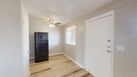 This single-floor unit is built with painted concrete block walls throughout, giving it a sturdy, low-maintenance character. Light wood-style laminate flooring runs continuously across the living areas and bedroom, creating a cohesive and clean aesthetic. The entry opens into a bright living area illuminated by a window with blinds, a ceiling fan with integrated lighting, and a textured ceiling. The kitchen is efficiently laid out and features gray cabinetry, white subway tile backsplash, light countertops, a double stainless steel sink, a black electric range, and an over-the-range exhaust hood. A full-size black refrigerator is positioned just outside the kitchen area. The bedroom is well-proportioned, receiving natural light through a sizable window, and includes access to a walk-in closet offering generous storage space. The bathroom is clean and straightforward, equipped with a single-sink vanity, a mirrored medicine cabinet, a toilet, and a tub-shower combination with tiled surround. Throughout the unit, white trim and doors provide a crisp contrast against the neutral-toned block walls, contributing to a tidy and functional overall appearance.