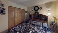 This single-floor home offers a collection of well-utilized living spaces designed for comfortable, everyday living. The residence includes multiple bedrooms, each with its own distinct character. One bedroom features a metal bunk bed with a full-size lower bunk, carpeted flooring, and a window with a light-filtering blind — ideal for shared sleeping arrangements. Another bedroom is outfitted with a full-size bed, a mirrored headboard, a sliding wood-panel closet, and decorative wall mirrors that add depth to the room. A third bedroom offers carpeted floors, a large double-door closet with classic six-panel wood doors, and a cozy bean bag chair nestled in the corner. A fourth bedroom features a window dressed in dark curtains, a lean mirror, and soft carpet underfoot. The bathroom is neatly finished with dark wood cabinetry, a granite-look countertop, an undermount sink, and framed mirrors accented by decorative seashell wall art, lending a coastal touch. Throughout the home, warm beige walls, carpeted floors, and wood-trimmed doors create a cohesive and inviting aesthetic. Windows in several rooms allow for natural light, contributing to the home's warm and welcoming ambiance.