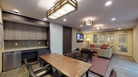 This thoughtfully designed single-floor space is organized into two distinct yet open zones: a dining area and a lounge area, unified by consistent warm-toned finishes and hardwood-style flooring. The dining area accommodates a large live-edge wood table surrounded by dark wood chairs, illuminated by a striking geometric brass-and-glass pendant light fixture. Large windows with horizontal blinds and a sliding glass door flood the area with natural light and provide direct access to a furnished outdoor patio, extending the usable space outdoors.

A sleek kitchenette, partially concealed by a full-height frosted sliding panel, features contemporary wood-grain cabinetry, a tile backsplash, an under-counter appliance, and a utility sink — ideal for catering or refreshment service.

The adjacent lounge area centers on a spacious sectional sofa in neutral tones accented with deep red throw pillows, arranged around a round wood coffee table atop a patterned area rug. A large flat-screen television is mounted on a textured stone accent wall, flanked by a contrasting dark painted wall, creating a bold focal point. The lounge also benefits from direct outdoor access through sliding glass doors. Recessed lighting and decorative ceiling-mounted fixtures provide layered illumination throughout.
