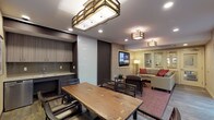 This thoughtfully designed single-floor space is organized into two distinct yet open zones: a dining area and a lounge area, unified by consistent warm-toned finishes and hardwood-style flooring. The dining area accommodates a large live-edge wood table surrounded by dark wood chairs, illuminated by a striking geometric brass-and-glass pendant light fixture. Large windows with horizontal blinds and a sliding glass door flood the area with natural light and provide direct access to a furnished outdoor patio, extending the usable space outdoors.

A sleek kitchenette, partially concealed by a full-height frosted sliding panel, features contemporary wood-grain cabinetry, a tile backsplash, an under-counter appliance, and a utility sink — ideal for catering or refreshment service.

The adjacent lounge area centers on a spacious sectional sofa in neutral tones accented with deep red throw pillows, arranged around a round wood coffee table atop a patterned area rug. A large flat-screen television is mounted on a textured stone accent wall, flanked by a contrasting dark painted wall, creating a bold focal point. The lounge also benefits from direct outdoor access through sliding glass doors. Recessed lighting and decorative ceiling-mounted fixtures provide layered illumination throughout.