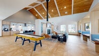 This expansive single-floor amenity space is thoughtfully designed to offer a comprehensive community experience across five distinct areas. At its heart lies an open-concept lounge and social hub defined by soaring vaulted ceilings with warm wood paneling and white exposed beams, creating a bright and airy atmosphere. The living area features a contemporary gas fireplace, a mounted flat-screen TV, plush seating, and a shuffleboard table, all set atop elegant herringbone-patterned hardwood flooring. Large windows frame outdoor views and flood the space with natural light.

The gourmet kitchen is a standout feature, showcasing a dramatic marble slab backsplash with custom-shaped cutouts, light wood cabinetry, a waterfall-edge marble island, and bar seating with woven leather chairs. A private dining nook offers built-in banquette seating in blue and cognac upholstery, a marble-topped table, and a picture window overlooking the exterior.

The fitness center is generously sized and well-equipped with treadmills, cycling bikes, strength machines, free weights, and functional training equipment, all beneath the same vaulted wood ceiling. A co-working lounge provides a quieter retreat with carpeted flooring and expansive windows overlooking a resort-style pool area, making it ideal for focused work or relaxation.