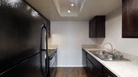 Located at 9400 Abercorn St in Savannah, Georgia, this single-floor space offers a practical and well-appointed layout. The kitchen is a standout feature, designed in a galley configuration that maximizes efficiency. It is outfitted with rich, dark espresso-toned cabinetry paired with light speckled countertops, creating a striking contrast throughout. The kitchen includes a full suite of black appliances — a refrigerator, a gas range, and a dishwasher — all neatly integrated along opposing walls. A stainless steel double-basin sink with a modern faucet is positioned on one side, while the opposite wall accommodates the range and refrigerator. The flooring throughout the kitchen is durable wood-look planking that adds warmth to the space. Above, a recessed tray ceiling detail with track lighting provides targeted illumination, enhancing both the functionality and aesthetic of the kitchen. The white walls keep the space feeling clean and open despite the galley layout. Overall, the space presents a cohesive, utilitarian design with thoughtful finishes suited for everyday living.