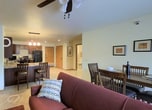 This single-floor space offers a welcoming and functional layout designed for comfortable everyday living. The living area serves as the heart of the home, featuring plush carpeting underfoot and warm, neutral wall tones that create a relaxed, inviting atmosphere. A pair of deep burgundy sofas are arranged around a dark wood coffee table, anchored by a rich, earth-toned area rug that adds texture and warmth to the room. Decorative accent pillows complement the color palette, lending a thoughtful, finished look to the seating arrangement. Natural light filters into the living area through a window dressed with vertical blinds, brightening the space and enhancing its open feel. A ceiling fan overhead provides both comfort and airflow. Tasteful wall art and ambient floor and table lamps contribute to the cozy, lived-in character of the room. A glimpse of a dining area is visible adjacent to the living space, suggesting a practical, open flow between shared living zones. Overall, the space presents a comfortable, no-fuss environment with a warm color scheme and functional design that makes it well-suited for relaxed, day-to-day living.