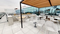 This sophisticated urban amenity space is designed to impress, offering a seamless blend of upscale interiors and expansive outdoor access across a single level. The open-plan lounge area is anchored by warm hardwood flooring, floor-to-ceiling glass walls, and generous natural light that frames panoramic city skyline views. A large marble-clad fireplace serves as a dramatic focal point, flanked by wood-panel wall accents and a mounted flat-screen display. Comfortable seating arrangements, including plush sofas, leather accent chairs, and intimate groupings, create versatile zones for socializing or relaxing.

A secondary lounge area near the glass perimeter offers additional seating with direct access to the rooftop terrace. The full kitchen features sleek white cabinetry, a bold marble-effect backsplash extending to the ceiling, a gas range with a stainless steel hood, a central island with bar seating, and pendant lighting — making it well-suited for catered events or private gatherings.

The rooftop terrace extends the space outdoors, featuring large-format tile flooring, a pergola with a warm wood-slat ceiling, white wicker and wood seating arrangements, raised planters, and unobstructed skyline views, creating a refined outdoor entertaining environment.