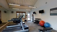 This fitness center offers a clean, functional workout environment designed to support a variety of exercise routines. The space is well-lit, benefiting from both natural light through a wood-framed window fitted with slatted blinds and bright overhead fluorescent strip lighting mounted to a white coffered ceiling, which lends a sense of openness to the room.

On one side of the facility, a row of Life Fitness treadmills provides dedicated space for cardio training. Opposite the cardio equipment, a tiered dumbbell rack holds a comprehensive range of free weights, complemented by a medicine ball rack stocked with multiple weighted balls and a stability ball for core training. A weight bench is also present, supporting strength-focused workouts.

Entertainment is provided by two wall-mounted flat-screen televisions, keeping users engaged during their sessions. The flooring features durable, gym-appropriate carpeting suited for high-traffic use. A wall-mounted fan adds to the ventilation of the space, while a fire extinguisher and emergency exit signage reflect attention to safety standards. A glass-paneled door provides direct access to the exterior, enhancing airflow and connectivity to the surrounding area. Overall, the facility is compact yet thoughtfully arranged to maximize usability for residents or guests.