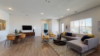 This well-appointed single-floor space combines modern design with functional versatility across several distinct areas. The expansive main living area features warm hardwood-style flooring, crisp white walls, and recessed lighting, creating a bright and welcoming atmosphere. A spacious lounge zone is anchored by a large sectional sofa, mustard accent chairs, and a geometric coffee table arranged atop a patterned area rug, with a wall-mounted flat-screen TV and a window offering natural light from outside.

Adjacent to the main lounge, a separate seating nook features deep charcoal-toned walls, tufted armchairs, navy blue ottomans, and distinctive teal sculptural wall art, giving the area a cozy, curated feel. A private meeting or conference room is accessible through sleek black-framed glass doors, furnished with a round table and amber-upholstered chairs, ideal for focused gatherings.

The kitchen is outfitted with rich dark cabinetry, granite countertops, stainless steel appliances including a French door refrigerator, a tile backsplash, and a generous island with bar seating. An entry area with bold orange accent chairs and clean white walls leads seamlessly into the broader space. Together, these zones create a cohesive, design-forward environment suited for both social and functional use.