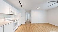 This single-floor unit efficiently accommodates four bedrooms and two bathrooms within a compact footprint, making it a highly functional multi-room layout. Light hardwood-style flooring runs consistently throughout the main living areas and bedrooms, contributing to a clean, cohesive aesthetic.

The kitchen features white cabinetry, white appliances including an electric range, over-the-range microwave, and refrigerator, along with track lighting overhead. It opens toward the living area, which is brightened by a window and equipped with a ceiling fan with integrated lighting. A second ceiling fan is also present in the primary bedroom, which receives natural light through a gridded window.

The remaining bedrooms are bright and simply finished, each with white walls, hardwood-style floors, and natural light from individual windows. One bedroom includes a built-in wardrobe unit for storage.

Both bathrooms maintain a consistent design language, featuring light wood-toned vanities with white countertops, large mirrors, walk-in shower stalls with adjustable handheld showerheads, and tile flooring. Two hallways connect the rooms, supporting a straightforward and navigable floor plan throughout.