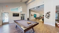 This generously sized, single-floor community amenity space is designed to accommodate a variety of social and recreational activities. The layout flows openly across distinct zones, each serving a specific function while maintaining a cohesive, modern aesthetic throughout.

The game room area is a prominent focal point, featuring a sleek, contemporary pool table set beneath a stylish pendant light fixture. Soaring vaulted ceilings in this section amplify the sense of openness, while large windows along the perimeter flood the space with natural light and offer views of the surrounding outdoor greenery. Wood-look flooring runs throughout, grounding the design with warmth and texture.

Adjacent to the game room, a well-appointed lounge area provides comfortable seating with tufted leather sectionals and mid-century modern accent chairs arranged around decorative area rugs. Bold teal artwork adds a vibrant pop of color against the neutral gray walls.

Further within the space, a casual dining and bar area features high-top tables and counter seating, complemented by patterned accent walls and a beverage station. A fitness area with exercise equipment is also accessible, visible through a mirrored wall section. Recessed lighting throughout ensures the entire space remains bright and inviting.