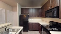This single-floor residence offers a thoughtfully designed layout with cohesive finishes throughout. The kitchen is a standout feature, outfitted with rich, dark espresso-toned cabinetry that extends from base to upper cabinets, providing generous storage. Light-toned countertops contrast the cabinetry, while black appliances — including a refrigerator, electric range, over-the-range microwave, and sink — complete the functional space. Light tile flooring runs through the kitchen, and a peninsula with a built-in sink adds additional workspace and separation from the living area. The open living area is finished with neutral-toned carpet and painted in warm beige tones. A ceiling fan with integrated lighting anchors the room, while multiple windows fitted with horizontal blinds allow natural light to filter in. Track lighting adds additional illumination options. The bathroom features a dark-toned vanity beneath a light countertop and single basin sink. A large mirror is positioned above the vanity, lit by a multi-bulb fixture above. A tiled tub and shower combination occupies one wall, completing the space. Overall, the unit presents a neutral, functional aesthetic with consistent design elements throughout.