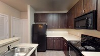This single-floor residence offers a thoughtfully designed layout with cohesive finishes throughout. The kitchen is a standout feature, outfitted with rich, dark espresso-toned cabinetry that extends from base to upper cabinets, providing generous storage. Light-toned countertops contrast the cabinetry, while black appliances — including a refrigerator, electric range, over-the-range microwave, and sink — complete the functional space. Light tile flooring runs through the kitchen, and a peninsula with a built-in sink adds additional workspace and separation from the living area. The open living area is finished with neutral-toned carpet and painted in warm beige tones. A ceiling fan with integrated lighting anchors the room, while multiple windows fitted with horizontal blinds allow natural light to filter in. Track lighting adds additional illumination options. The bathroom features a dark-toned vanity beneath a light countertop and single basin sink. A large mirror is positioned above the vanity, lit by a multi-bulb fixture above. A tiled tub and shower combination occupies one wall, completing the space. Overall, the unit presents a neutral, functional aesthetic with consistent design elements throughout.