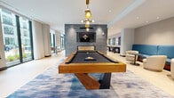 Located at 74 Freeman St in Newark, NJ, this contemporary community lounge offers a generously sized, open-plan living area designed with modern sophistication. The space is anchored by a striking textured stone accent wall that houses a wall-mounted TV and a sleek linear gas fireplace, creating a dramatic focal point throughout the room. A premium billiards table with a solid wood frame sits atop an abstract blue area rug and is illuminated by a sculptural brass chandelier, lending the space a polished, upscale atmosphere. Adjacent to the game area, a comfortable lounge features a rich cognac leather sofa, velvet ottomans, and a glass-and-gold coffee table, positioned to enjoy both the fireplace and natural light. A bar counter with deep blue tile backsplash, warm wood cabinetry, and navy velvet bar stools serves as a stylish social hub, accented by pendant lighting. Floor-to-ceiling windows with sheer white drapery flood the interior with natural light and offer open courtyard views, while light hardwood flooring ties the entire space together. Additional seating nooks and lounge chairs are distributed throughout, making this an inviting, multi-use gathering space ideal for socializing and recreation.