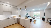 This contemporary 2-floor residence showcases a thoughtfully designed open-concept layout that seamlessly connects the kitchen, dining, and living areas. The kitchen is outfitted with sleek gray flat-front cabinetry, white subway tile backsplash, quartz countertops, stainless steel appliances, and a central island with bar seating — all illuminated by elegant brass pendant lights. Light hardwood flooring flows throughout, enhancing the bright, airy atmosphere created by recessed lighting and an abundance of natural light. The open living area offers a comfortable, inviting atmosphere with direct access to an outdoor patio through large sliding glass doors, bringing in lush greenery views. The bathrooms are finished to a high standard, featuring floating white vanities with quartz counters, undermount sinks, chrome fixtures, and stylish oval and round black-framed mirrors. One bathroom includes a separate glass-enclosed shower with tile surround, while another features a clean, modern aesthetic with bold circular wall art. Recessed lighting and wall sconces add warmth and functionality throughout. The overall design palette — crisp whites, soft grays, warm wood tones, and brass accents — creates a cohesive, modern aesthetic that is both refined and welcoming across both levels of the home.