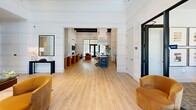 Located at 4170-4190 E Commerce Way in Sacramento, CA, this single-story clubhouse offers a well-appointed and thoughtfully designed communal space. Upon entry, a bright reception area greets visitors with soaring ceilings, floor-to-ceiling windows that invite abundant natural light, a glass-topped desk, and plush gold velvet accent chairs. A sleek credenza with decorative brass accents lines the wall, setting an upscale tone throughout. The open main lounge area features light wood flooring, horizontal shiplap-style wall paneling, and exposed beam ceiling details that lend architectural character. A built-in fireplace serves as the focal point, flanked by a mounted television and floating shelves with curated décor. Comfortable seating arrangements with swivel chairs, sofas, and mixed-material accent tables create an inviting gathering atmosphere. Adjacent to the lounge, a dining area features a substantial dark wood communal table surrounded by rounded cream upholstered chairs, large-scale artwork, and direct access to an outdoor patio. A private meeting nook offers a round glass table, gold velvet chairs, and a sputnik-style pendant light, while a well-lit corridor with black-framed glass doors connects the various areas of the space seamlessly. A beverage station and kitchenette add practical convenience throughout.