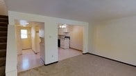 This two-floor space features an open-concept layout that connects the carpeted living area to the kitchen, creating a cohesive and functional flow. The living room is finished with neutral-toned carpet and light-colored walls, contributing to a clean, understated aesthetic. A wide, open passageway leads directly into the kitchen, which features vinyl flooring, cream-colored cabinetry, and a white electric range with an overhead cabinet for added storage. A ceiling fan with a light fixture illuminates the kitchen area, providing both ventilation and practical lighting. The kitchen walls feature a subtle striped wallpaper accent, adding a touch of texture to the space. A hallway visible from the living area suggests additional rooms or access points within the layout. The overall color palette of whites, creams, and soft neutrals gives the space a bright and airy feel. With its straightforward design and practical features spread across two floors, this space offers a functional and comfortable environment suited for everyday living.
