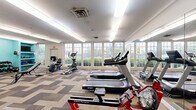 This fitness center offers a comprehensive exercise space thoughtfully designed to accommodate a variety of workout routines. The room is anchored by a bank of large, multi-pane windows along one wall, flooding the interior with abundant natural light and providing pleasant views of lush greenery outside. A full-length wall mirror runs along one side, visually expanding the space and allowing users to monitor their form during workouts.

The cardio section is well-stocked with multiple Spirit treadmills, an upright elliptical trainer, a recumbent bike, and a rowing machine, providing ample options for aerobic exercise. Toward the back of the room, a vibrant teal accent wall distinguishes the strength training area, which features a multi-station cable machine, a rack of dumbbells in graduated weights, a weight bench, and an open shelving unit holding kettlebells, resistance bands, and exercise mats. A wall-mounted television is conveniently positioned in this area. A vending machine near the entrance adds a practical amenity for users. The flooring throughout consists of a patterned, multi-tone carpet tile in neutral tones of beige, gray, and brown. Overhead fluorescent lighting supplements the natural light, ensuring the space remains bright and functional throughout the day.