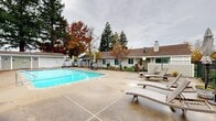 Located at 2529 Eastern Ave in the Arden-Arcade neighborhood of Sacramento, CA, this two-story residential community offers a well-appointed shared outdoor environment complemented by thoughtfully maintained grounds. The complex is organized across multiple numbered buildings — visible signage identifies individual unit clusters — each constructed with board-and-batten siding in a neutral gray palette beneath pitched rooflines, lending the property a cohesive, understated aesthetic.

At the heart of the community lies a generously sized outdoor swimming pool, surrounded by a spacious concrete deck ideal for lounging. Rattan-style chaise lounges are positioned poolside, offering residents a comfortable setting for relaxation. The pool area is enclosed by a dark metal iron fence that provides both security and visual definition throughout the grounds.

Adjacent to the pool, a dedicated amenity building houses accessible restroom and shower facilities, clearly marked for both men and women, with ADA-compliant access. An outdoor rinse shower is also available at poolside.

Mature trees — including towering pines and deciduous specimens displaying seasonal color — frame the property and provide natural shade and privacy. Manicured hedgerows line the walkways between buildings, adding greenery and structure to the landscape. Globe-style exterior lighting fixtures are positioned throughout, ensuring the community remains well-lit after dark.