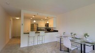 This single-floor residence showcases a clean, modern aesthetic throughout its open-concept layout. The kitchen is a standout feature, outfitted with crisp white cabinetry, dark granite countertops, and stainless steel appliances including a full-size refrigerator and built-in microwave. Recessed lighting is complemented by elegant pendant lights that hang above the breakfast bar, creating a warm and inviting atmosphere. The breakfast bar, finished with the same dark granite, provides casual seating with stylish chrome and woven bar stools, making it an ideal spot for quick meals or entertaining guests. Adjacent to the kitchen, the dining area offers additional seating around a glass-top table paired with matching white woven chairs, maintaining a cohesive and contemporary design language. The living and dining spaces feature neutral-toned carpeting that adds a soft, comfortable feel underfoot. The open layout between the kitchen, dining, and living areas creates a seamless flow, maximizing the sense of space. The neutral color palette throughout — white walls, white cabinetry, and light flooring — contributes to a bright, airy ambiance that feels both spacious and welcoming.