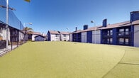 Located at 9669 Forest Ln in Dallas, Texas, this outdoor recreational space is a well-appointed, single-level facility centered around a large, open artificial turf field. The vibrant green synthetic surface provides a low-maintenance, all-weather playing area suitable for a variety of sports and recreational activities. The field is fully enclosed by sturdy chain-link fencing along the perimeter, complemented by tall overhead netting that contains balls and ensures a safe, contained playing environment. Tall light poles are positioned throughout the space, indicating the field is equipped for evening use. The surrounding multi-story buildings feature white and navy blue exteriors with balconies, stairwells, and pitched rooftops with chimneys, forming a courtyard-like setting around the turf area. A glimpse of a pool area with covered seating is visible beyond the fencing on one side, suggesting the turf field is part of a broader amenity complex. The open-air design allows for abundant natural light and clear sky views, creating a bright and energetic atmosphere. The space is well-suited for organized sports, casual recreation, or community gatherings, offering a functional and prominent outdoor amenity within the surrounding residential community.