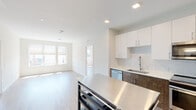 This contemporary single-floor unit is designed with a clean, modern aesthetic characterized by crisp white walls, light hardwood flooring, and an abundance of natural light throughout. The open-concept layout seamlessly connects the kitchen and living area, creating a bright and functional living space.

The kitchen is a standout feature, outfitted with a two-tone cabinet design combining white upper cabinetry and rich dark wood lower cabinets, accented by a classic white subway tile backsplash. Stainless steel appliances include a refrigerator, electric range, over-the-range microwave, and dishwasher. A stainless steel kitchen island provides additional workspace and storage. Recessed lighting illuminates the area efficiently.

The bedrooms are generously sized, each featuring large windows that invite natural light and views of the surrounding area, along with light hardwood flooring and neutral walls. The layout, as shown in the 3D overview, accommodates multiple bedrooms connected by a central corridor.

Two bathrooms complete the unit. One features a glass-enclosed walk-in shower with floor-to-ceiling white tile and chrome fixtures. The second includes a wood-toned vanity with a white countertop and a large mirror. Both bathrooms feature light tile flooring, maintaining the unit's cohesive modern palette throughout.
