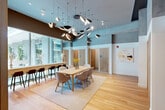 Located at 77 Bruton St in San Francisco, CA, this single-floor space is a well-appointed shared workspace designed to foster both focused work and collaboration. The heart of the space is a bright, open common area anchored by a large communal dining-style table with modern upholstered chairs, set atop a patterned area rug over warm hardwood flooring. Soaring ceilings create a dramatic sense of volume, further accentuated by a sculptural chandelier featuring asymmetric pendant light fixtures that serve as a striking artistic focal point overhead. The walls are finished in a two-tone treatment of deep blue-gray above and clean white below, lending the space a contemporary and refined ambiance. Floor-to-ceiling glass windows line one side of the room, flooding the interior with abundant natural light and offering views of an adjacent courtyard with greenery. A bar-height counter with dark stools runs along the window wall, providing additional workstation options. Multiple private rooms — including dedicated office and bedroom spaces — are accessible via elegant slatted wood-paneled dividers, each identified with letter signage. An entrance and hallway connect the various zones seamlessly. Abstract artwork adorns the walls, adding a curated, gallery-like quality to the overall environment.