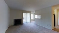 This single-floor residence offers a functional and straightforward layout designed for comfortable everyday living. The living room is finished with wall-to-wall carpeting and neutral-toned walls, creating a clean, understated atmosphere. A tile-surround fireplace anchors one wall, providing a focal point and a source of warmth. A full-length sliding glass door fitted with vertical blinds allows natural light to filter into the space and offers access to the outdoors. A pass-through window connects the living area to the kitchen, enhancing the sense of openness between the two spaces. An additional window brings in further natural light.

The galley-style kitchen is efficiently organized, with upper and lower cabinetry lining both sides of the corridor layout. Wood-toned upper cabinets provide generous storage, while the countertops offer practical workspace. Appliances include a stainless steel refrigerator, a black electric range, and a dishwasher. A single-basin sink is positioned beneath the upper cabinets on one side. Dark tile flooring runs throughout the kitchen, providing a durable and easy-to-maintain surface. A hallway at the end of the kitchen leads to additional areas of the unit.