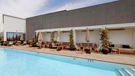 This rooftop pool deck is a refined outdoor retreat designed for both relaxation and social gathering. At its centerpiece is a generously sized lap pool with crystal-clear turquoise water, flanked by teak chaise loungers outfitted with bold striped cushions. A row of semi-enclosed cabanas lines one side of the pool, each furnished with plush sofas, accent chairs, and side tables beneath white pergola-style canopies, offering shaded privacy while remaining open to the outdoors. Climbing greenery and tall potted plants frame the cabanas, adding a lush, organic quality to the space.

The deck is paved in large-format neutral stone tiles that lend a clean, cohesive aesthetic throughout. Scattered potted succulents, palms, and mature trees punctuate the space, softening the architectural elements. Floor-to-ceiling glass doors provide seamless indoor-outdoor access. The perimeter opens to sweeping panoramic views of the surrounding cityscape, rolling hills, and distant skyline, framed by sleek glass railings. Patio heaters positioned throughout the deck extend usability across seasons, making this a versatile, upscale outdoor amenity.