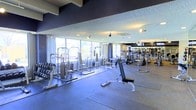 This modern fitness center offers a thoughtfully designed, open workout environment suited for a full range of exercise routines. The space is anchored by large floor-to-ceiling windows along one wall, flooding the interior with natural light and providing views of the surrounding greenery — creating an inviting atmosphere for training. The flooring features durable, dark rubberized material ideal for both free weight and machine use.

The strength training area is well-appointed with a wide selection of dumbbells neatly organized on a dedicated rack, alongside multiple cable machines, adjustable benches, and multi-function strength stations that accommodate a variety of exercises. The layout is spacious, allowing ample room between equipment for safe and comfortable movement.

Toward the rear of the facility, a dedicated cardio zone houses a row of treadmills and additional cardiovascular equipment, complemented by wall-mounted screens for entertainment during workouts. Track lighting and recessed ceiling fixtures provide consistent, even illumination throughout the space. Full-length mirrors line sections of the walls, enhancing the sense of openness while allowing users to monitor their form. The overall ambiance is clean, functional, and professional, making this fitness center a well-rounded facility for all fitness levels.