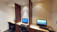 This single-floor business center is thoughtfully designed to accommodate productivity and focused work. The space features multiple individual computer workstations, each equipped with a desktop monitor, keyboard, and mouse, set into built-in desks with under-desk drawer units for convenient storage. Rich dark wood partitions divide the workstations, providing users with a degree of privacy while maintaining an open, collaborative atmosphere.

Recessed ceiling lighting illuminates each workstation with warm, directed light, creating a comfortable and functional environment. The neutral cream and beige wall tones contribute to a calm, professional ambiance throughout.

A standout architectural feature is a striking shoji-inspired partition wall composed of a wood-framed grid filled with frosted panels, flanking a dark wood glass-paned door. This elegant design element adds visual interest and allows diffused light to pass through, enhancing the sense of openness.

The space also includes a dedicated multifunction printer and scanner station, making it fully equipped for document handling needs. A small bookshelf with reference materials and a bulletin board round out the amenities, adding a practical and welcoming touch. The carpeted flooring throughout contributes to a quiet, focused working environment, making this business center a comprehensive and professional facility.