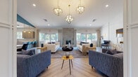 Located at 89 Privilege Rd in Bloomfield, Connecticut, this single-floor community space combines sophisticated design with functional amenities. The centerpiece is a generously proportioned open lounge area finished with light herringbone-patterned hardwood floors, crisp white walls with decorative paneling, and soaring ceilings adorned with striking gold orb chandeliers. A stone-clad fireplace anchors the main seating area, which is surrounded by an arrangement of plush sofas and accent chairs in coordinating blue and neutral tones. Expansive windows line the perimeter, flooding the interior with abundant natural light and offering views of the surrounding greenery. A dedicated media lounge area features elegant wainscoting, velvet accent chairs, wall sconces, and a large flat-screen display, creating an intimate yet refined setting. The well-appointed kitchen includes custom flat-panel cabinetry in a warm taupe finish, pristine white quartz countertops, a textured mosaic tile backsplash, stainless steel appliances, and generous counter space — designed to support both everyday use and group gatherings. Throughout the space, gold-toned fixtures and carefully curated décor provide a cohesive, upscale aesthetic. The overall ambiance is welcoming and polished, balancing comfort and contemporary style.